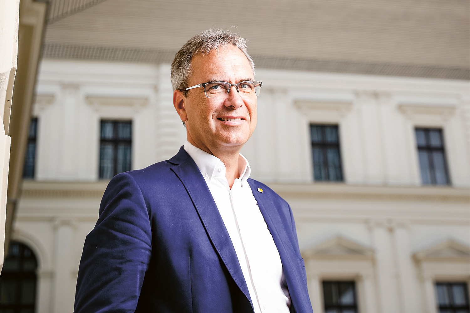 Ein Mann in einem blauen Anzug und weißem Hemd steht vor einem hellen Gebäude mit großen Fenstern und lächelt in die Kamera. Die Beleuchtung ist hell und natürlich.