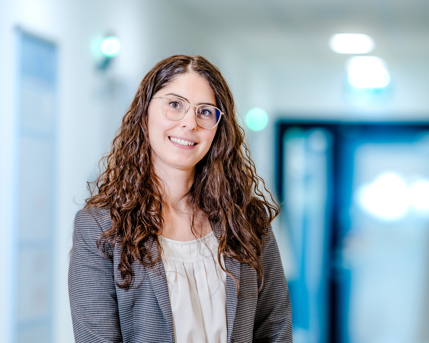 Eine Frau mit langen lockigen braunen Haaren, die eine Brille, einen grauen Blazer und eine cremefarbene Bluse trägt, lächelt, während sie in einem hell erleuchteten Flur mit blauen Akzenten steht.