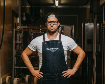 Ein Mann mit Brille und Mütze trägt eine dunkle Jeansschürze mit der Aufschrift "Buchberger" über einem weißen T-Shirt und steht mit den Händen in den Hüften in einer schwach beleuchteten Industrie- oder Werkstattumgebung.