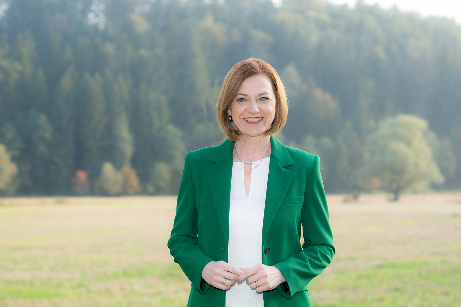 Eine Frau mit schulterlangen braunen Haaren, die einen grünen Blazer über einem weißen Oberteil trägt, steht an einem bewölkten Tag lächelnd auf einem Feld mit Bäumen und Grünpflanzen im Hintergrund.