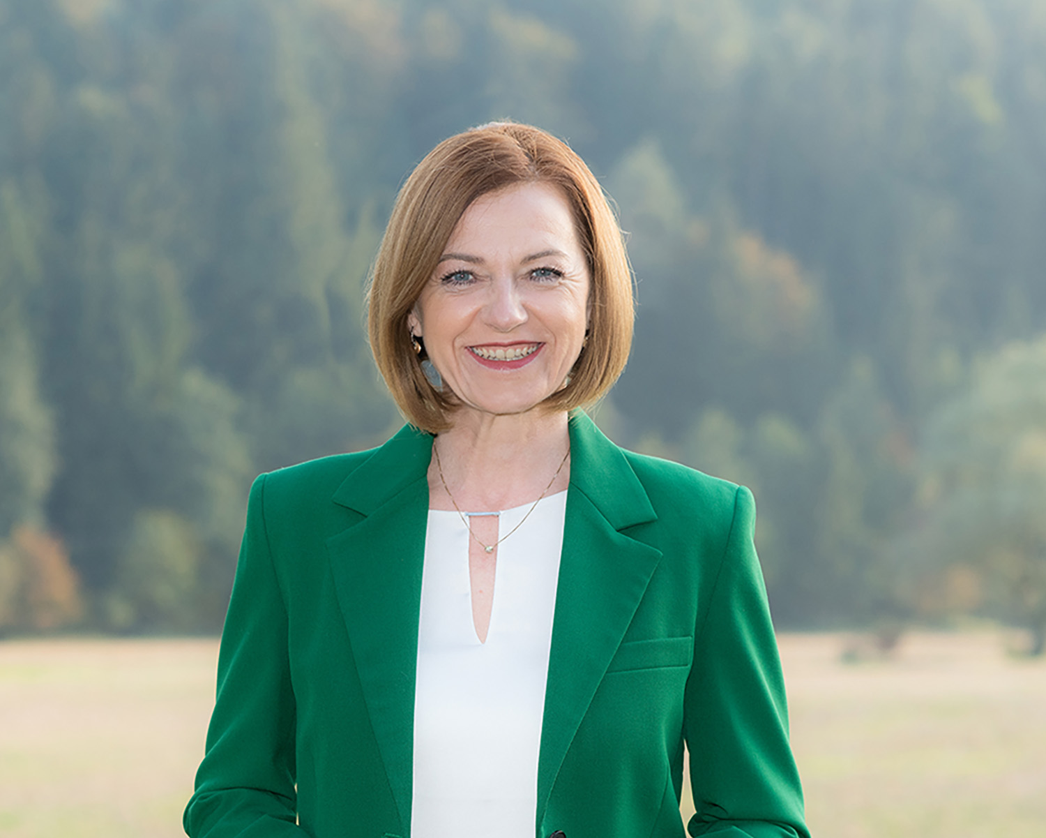 Eine Frau mit glattem, schulterlangem hellbraunem Haar, die einen grünen Blazer und ein weißes Oberteil trägt, lächelt, während sie im Freien vor einem verschwommenen Hintergrund aus Bäumen und Gras steht.
