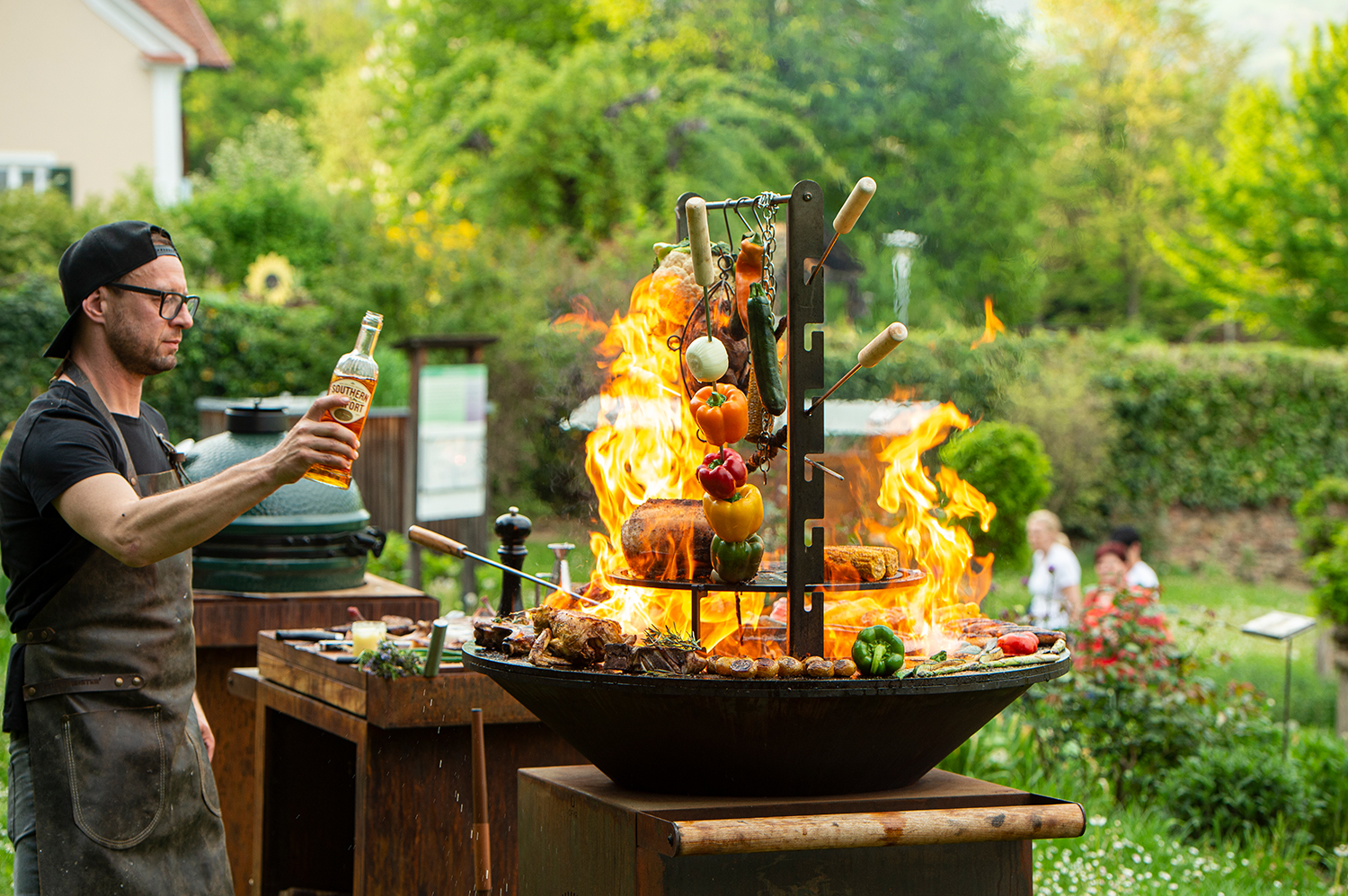 Ein Mann mit Brille und Schürze brät Fleisch und Gemüse über einer offenen Flamme auf einem Grill im Freien, in der einen Hand eine Flasche. Die Szene spielt in einem üppigen, grünen Garten.
