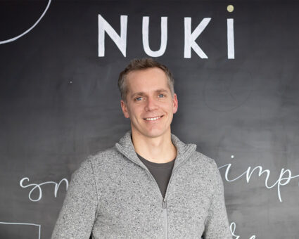 Ein Mann mit kurzen grauen Haaren und einem grauen Pullover mit Reißverschluss steht lächelnd vor einer Tafel, auf der das Wort "NUKI" steht.