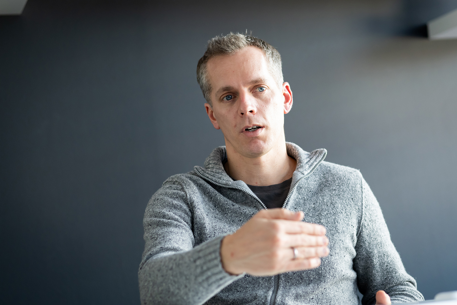 Ein Mann in einem grauen Pullover gestikuliert mit seiner rechten Hand, während er vor einem schlichten, dunklen Hintergrund sitzt und spricht. Er hat kurze Haare und einen ernsten Gesichtsausdruck.