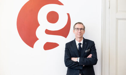Ein Mann in einem dunklen Anzug und mit Brille steht mit verschränkten Armen vor einer weißen Wand, auf der ein großer roter Kreis mit einem kleinen "g" zu sehen ist.