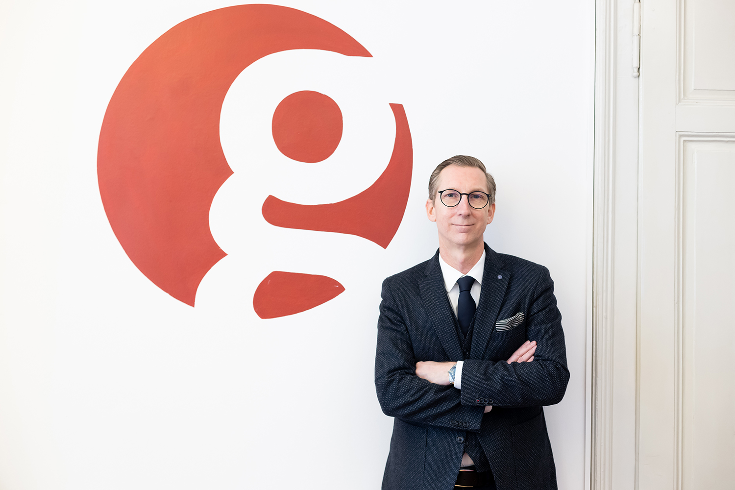 Ein Mann in einem dunklen Anzug und mit Brille steht mit verschränkten Armen vor einer weißen Wand, auf der ein großer roter Kreis mit einem kleinen "g" zu sehen ist.