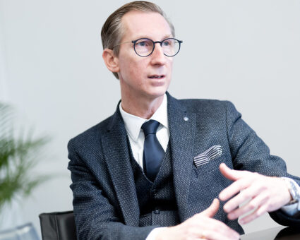 Ein Mann in einem dunklen Anzug, weißem Hemd und Krawatte sitzt in einem Haus und gestikuliert mit seinen Händen, während er spricht. Er hat kurzes hellbraunes Haar, trägt eine runde Brille und scheint in ein Gespräch vertieft zu sein.