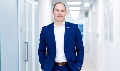 Ein Mann in einem blauen Anzug mit weißem Hemd steht in einem hellen, modernen Flur, die Hände in den Taschen und lächelt in die Kamera. Der Korridor hat Glastüren und gerahmte Bilder an den Wänden.