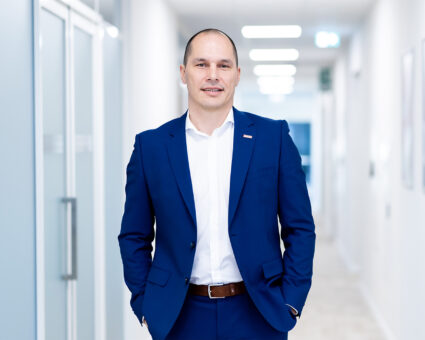 Ein Mann in blauem Anzug und weißem Hemd steht in einem hellen, modernen Flur, hat die Hände in den Taschen und lächelt selbstbewusst in die Kamera.