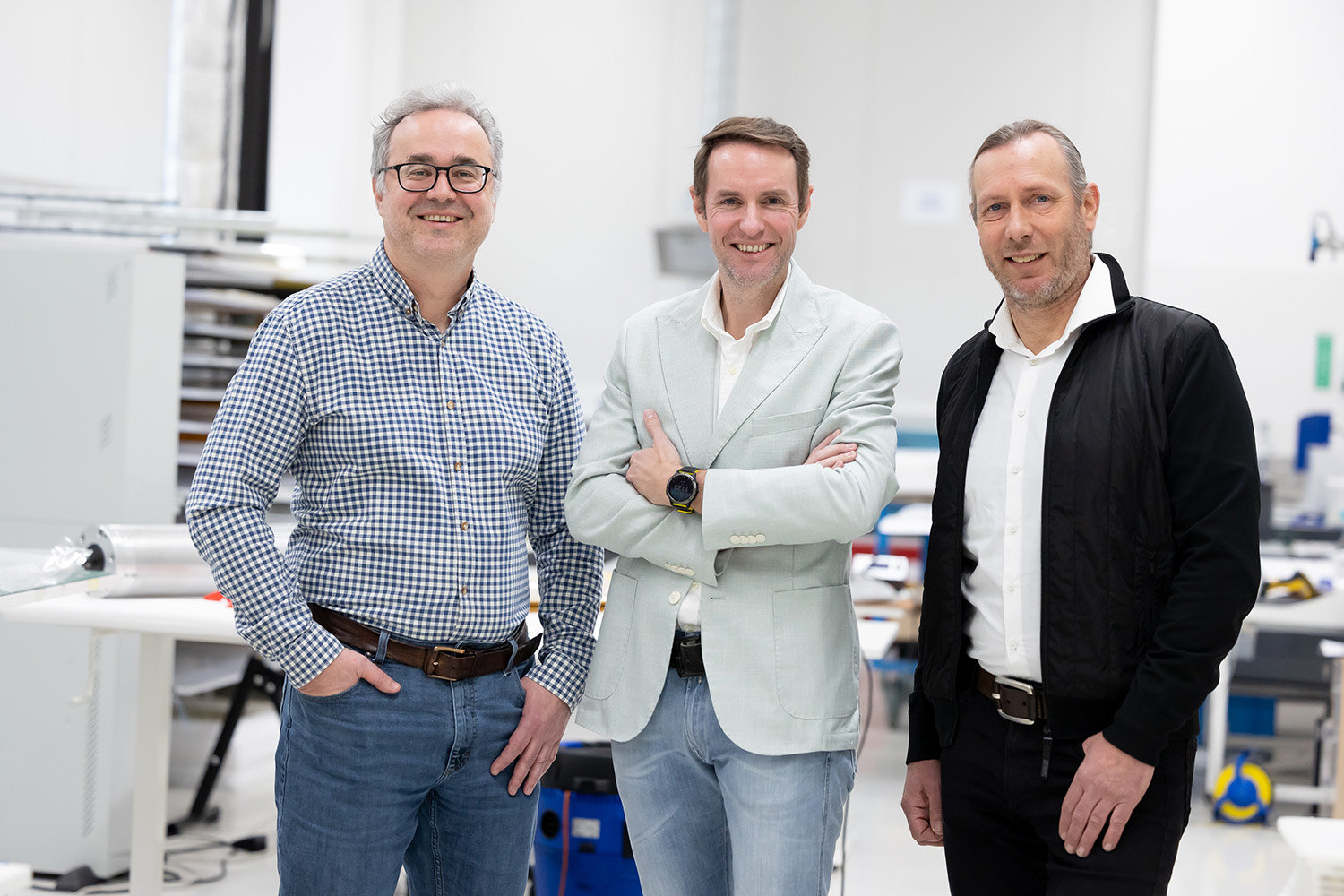Drei Männer stehen drinnen in einem hellen, modernen Büro oder Laborraum. Sie lächeln in die Kamera. Der Mann auf der linken Seite trägt ein kariertes Hemd, der Mann in der Mitte einen hellen Blazer und der Mann auf der rechten Seite eine schwarze Jacke.
