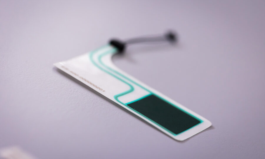 Nahaufnahme eines dünnen, flexiblen elektronischen Sensors mit Schaltkreismustern und einem kleinen schwarzen Bauteil auf einer glatten, hellgrauen Oberfläche.