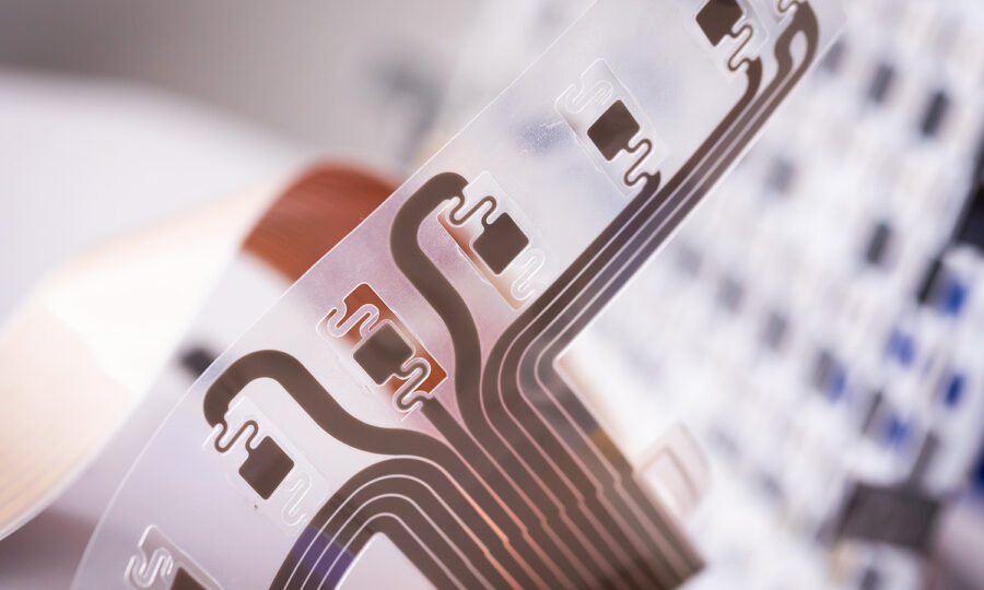 Nahaufnahme eines flexiblen, transparenten elektronischen Schaltkreises mit dunklen Leiterbahnen und chipähnlichen Komponenten, die vor einem unscharfen Hintergrund gehalten werden, der eine Tastatur oder ein elektronisches Gerät zu sein scheint.