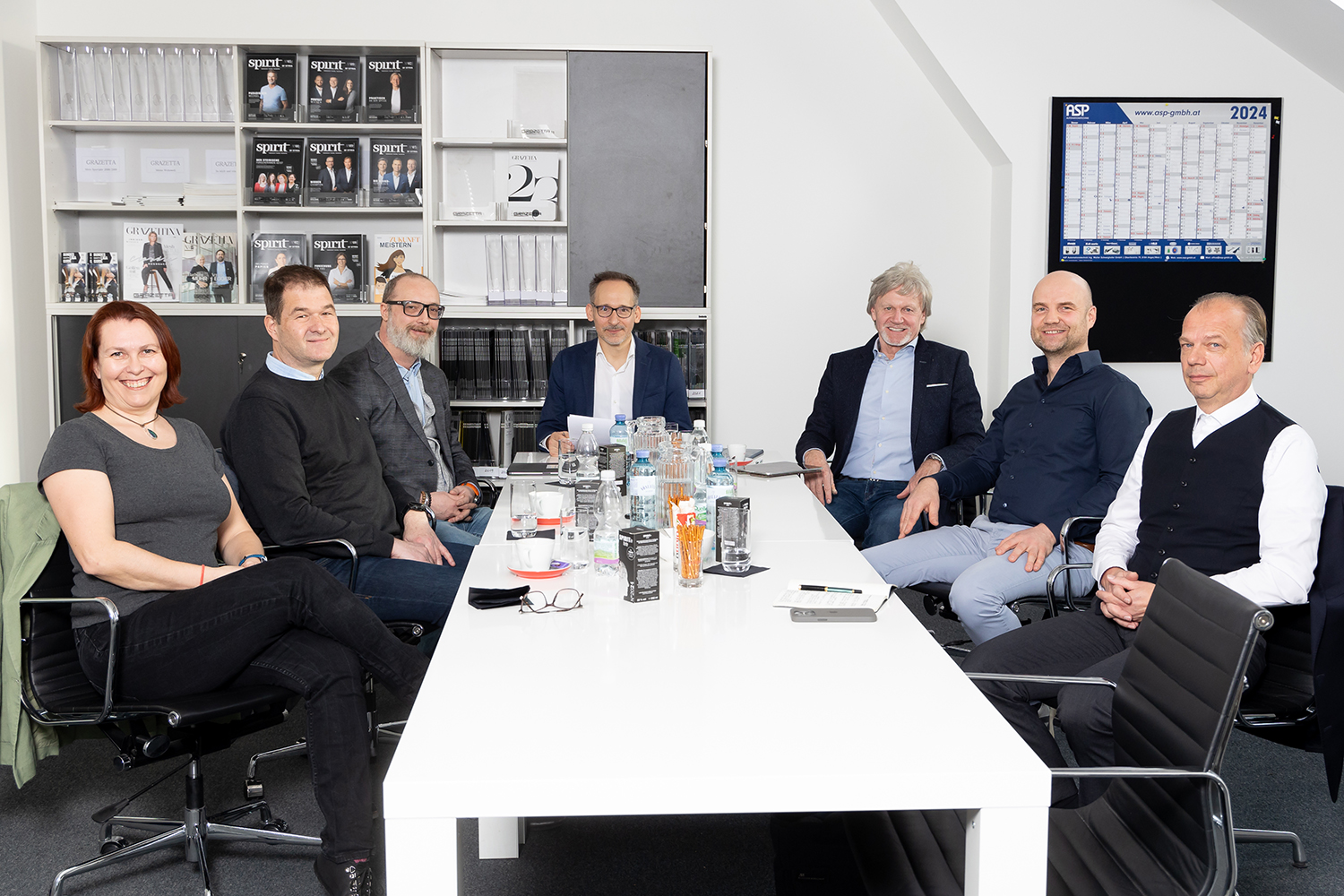 Sieben Personen, sechs Männer und eine Frau, sitzen um einen weißen Konferenztisch in einem Bürobesprechungsraum. Auf dem Tisch stehen Getränke, Papiere und Snacks. Im Hintergrund sind Regale mit Zeitschriften und ein Wandkalender zu sehen.