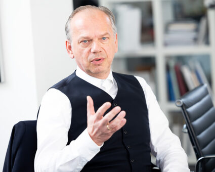 Ein Mann mittleren Alters mit kurzen grauen Haaren, der ein weißes Hemd und eine schwarze Weste trägt, gestikuliert mit seiner rechten Hand, während er in einem Büro sitzt, in dem Regale und Bücher im Hintergrund zu sehen sind.