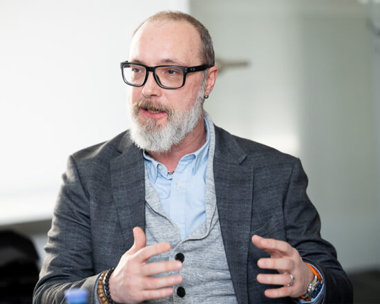 Ein Mann mit Brille und grauem Bart, der einen grauen Blazer über einem hellblauen Hemd trägt, gestikuliert mit seinen Händen, während er in einem hell erleuchteten Innenraum spricht.