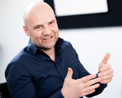 Ein lächelnder, glatzköpfiger Mann in einem dunkelblauen Hemd gestikuliert mit seinen Händen, während er in einem Haus sitzt und engagiert und ansprechbar wirkt. Der Hintergrund ist unscharf und hell.