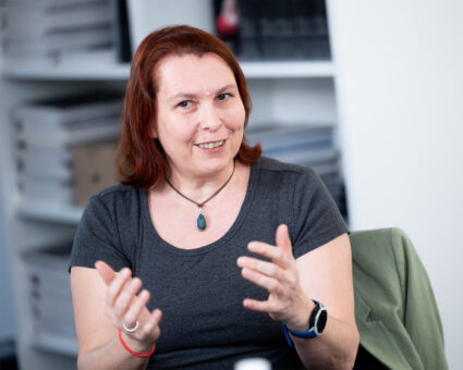 Eine Frau mit schulterlangen rotbraunen Haaren, die ein graues Hemd, eine türkisfarbene Halskette mit Anhänger und eine Smartwatch trägt, gestikuliert mit ihren Händen und lächelt in einem Büro mit Regalen im Hintergrund.