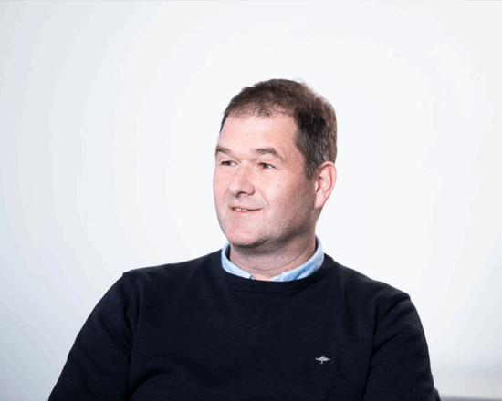 Ein Mann mittleren Alters mit kurzen braunen Haaren, der einen dunklen Pullover über einem hellblauen Hemd mit Kragen trägt, sitzt und lächelt, während er vor einem schlichten weißen Hintergrund leicht zur Seite blickt.