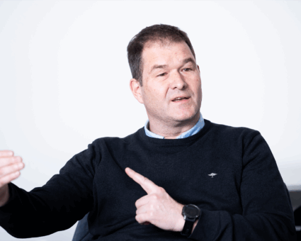 Ein Mann mit kurzen dunklen Haaren, der einen schwarzen Pullover und eine Uhr trägt, gestikuliert mit seiner rechten Hand, während er spricht. Er scheint sich vor einem schlichten, hellen Hintergrund zu unterhalten.