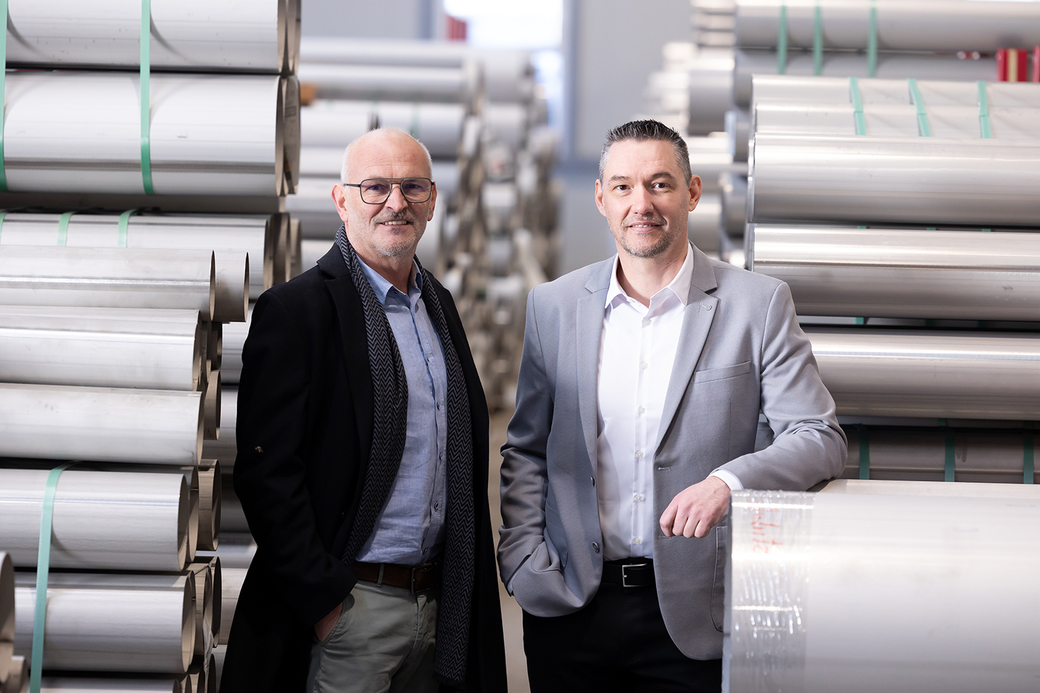 Zwei Männer in Geschäftskleidung stehen selbstbewusst in einem Lagerhaus, das mit großen, in Reihen gestapelten Rollen von Metallblechen gefüllt ist. Der Hintergrund zeigt eine industrielle Umgebung mit metallischen Materialien.