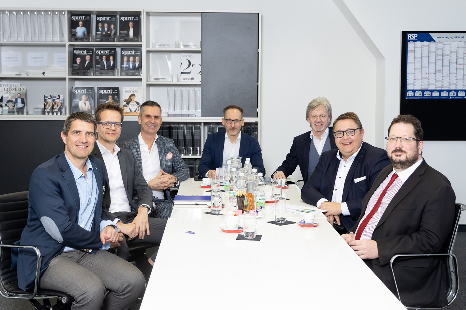 Sieben Männer in Geschäftskleidung sitzen um einen Konferenztisch mit Getränken und Papieren und lächeln in die Kamera. Im Hintergrund sind Regale mit Zeitschriften und Büchern sowie ein großer Kalender zu sehen.