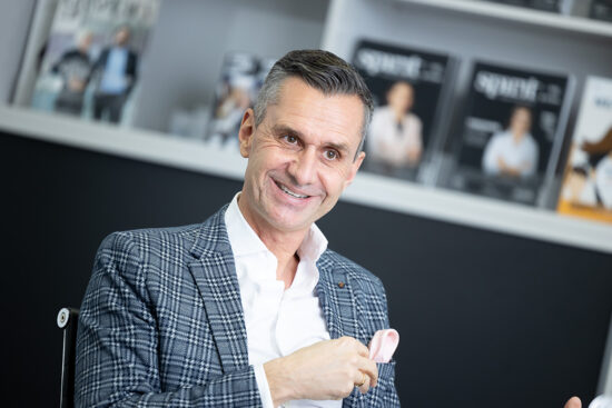 Ein lächelnder Mann in einem karierten Blazer sitzt in einem Haus und hält ein rosa Einstecktuch. In den Regalen im Hintergrund liegen unscharfe Zeitschriften aus.