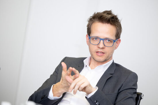 Ein Mann in einem dunklen Anzug und mit einer blauen Brille sitzt und spricht und gestikuliert mit seinen Händen. Der Hintergrund ist schlicht und unscharf.