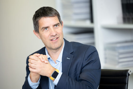 Ein Mann in einer blauen Anzugsjacke sitzt in einem Büro, faltet die Hände und lächelt leicht. Er hat kurzes dunkles Haar und trägt eine Smartwatch. In den Regalen im Hintergrund sind Stapel von Dokumenten zu sehen.