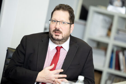 Ein Mann im schwarzen Anzug, mit roter Krawatte und Brille sitzt in einem Haus und spricht mit einer Hand auf der Brust. Im unscharfen Hintergrund sind Regale mit Büchern und Akten zu sehen.