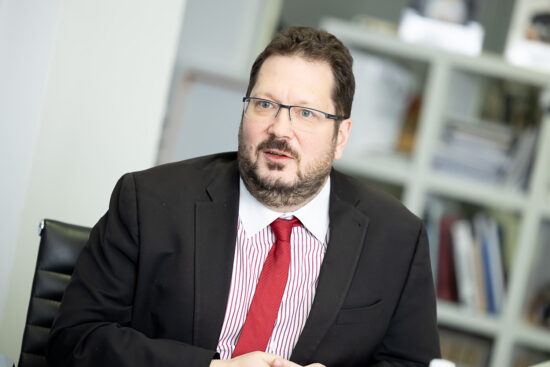 Ein Mann mit Brille, schwarzem Anzug, roter Krawatte und gestreiftem Hemd sitzt an einem Tisch in einem Büro mit Regalen und Büchern im Hintergrund und schaut leicht zur Seite.