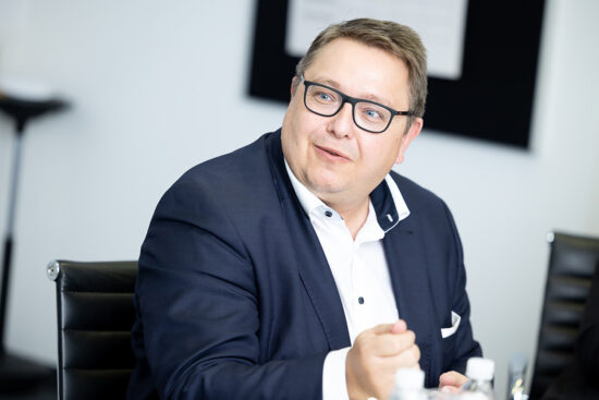 Ein Mann in einem marineblauen Anzug und mit Brille sitzt an einem Tisch, schaut zur Seite und lächelt. Er hält einen Stift in der Hand und scheint sich in einer hellen Büroumgebung zu unterhalten.