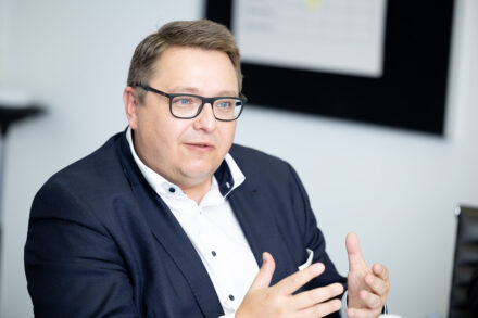 Ein Mann, der eine Brille, einen dunklen Anzug und ein weißes Hemd trägt, spricht und gestikuliert mit seinen Händen während einer Besprechung in einer hellen Büroumgebung.