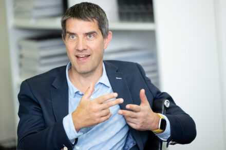 Ein Mann in einem blauen Blazer und hellblauem Hemd spricht angeregt und gestikuliert mit den Händen. Er hat kurzes dunkles Haar und sitzt in einem Raum, in dem im Hintergrund Bücherregale zu sehen sind.