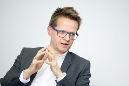 Ein Mann in einem grauen Anzug und mit einer blauen Brille gestikuliert mit seinen Händen, während er vor einem schlichten hellen Hintergrund spricht.