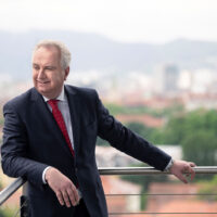 Ein Mann in einem dunklen Anzug und mit roter Krawatte steht im Freien und stützt sich auf ein Metallgeländer, mit einer verschwommenen Stadtlandschaft und Hügeln im Hintergrund. Er lächelt und blickt zur Seite.