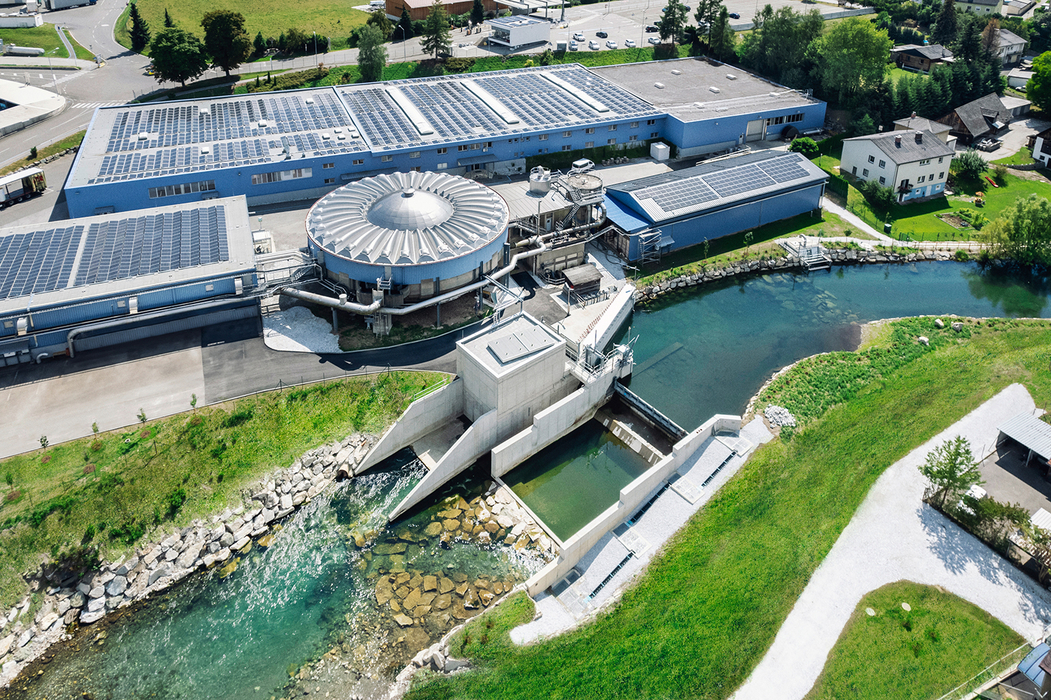 Luftaufnahme einer großen industriellen Wasseraufbereitungsanlage mit runden und rechteckigen Gebäuden, Sonnenkollektoren auf den Dächern und Wasser, das durch Kanäle in einen klaren Fluss fließt, der von grünem Gras und Straßen umgeben ist.
