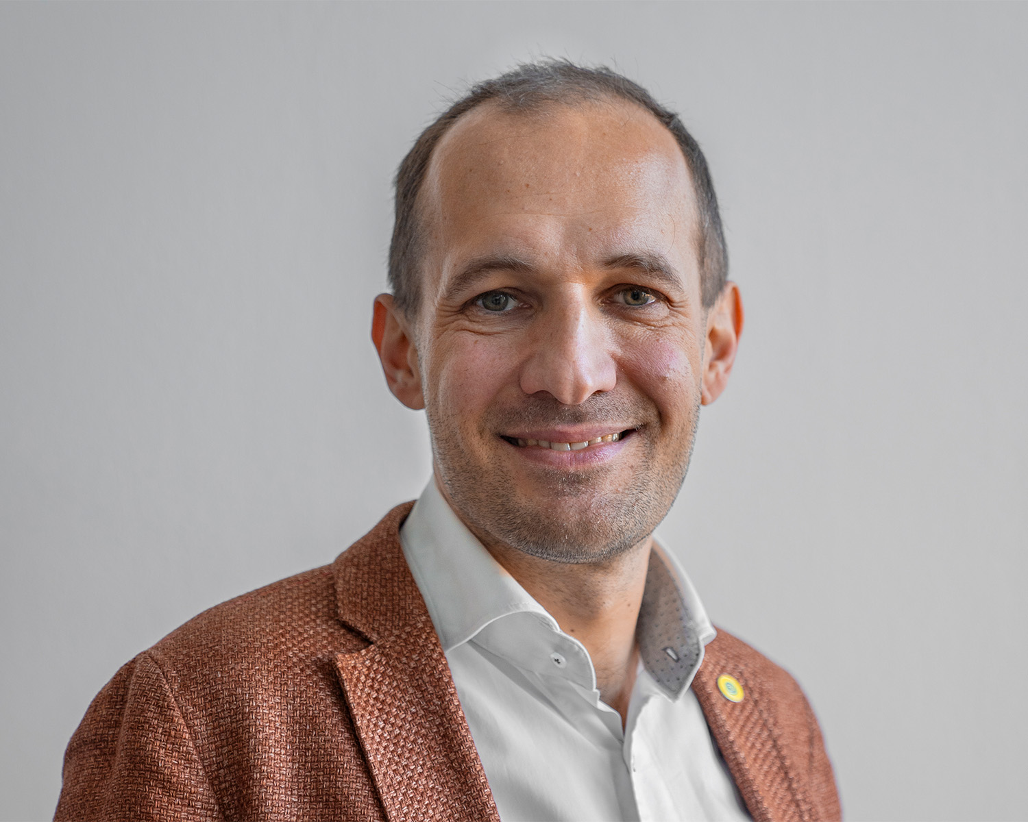Ein lächelnder Mann mit kurzen braunen Haaren trägt einen braunen strukturierten Blazer über einem weißen Hemd und steht vor einem schlichten hellgrauen Hintergrund.