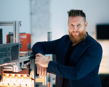 Ein Mann mit rotem Bart und Schnurrbart, der einen marineblauen Blazer trägt, steht selbstbewusst neben einer industriellen Ausrüstung in einem modernen, gut beleuchteten Labor oder einer Werkstatt.