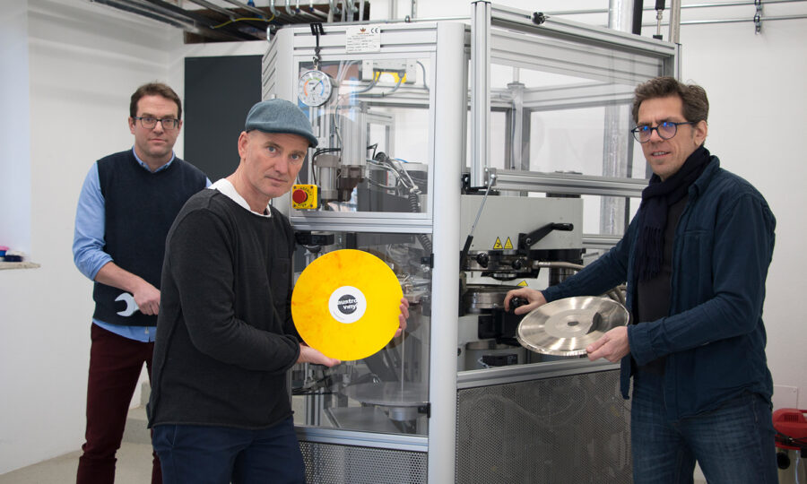 Drei Männer stehen vor einer Maschine, die Schallplatten presst. Einer hält eine gelbe Schallplatte, ein anderer eine silberne Schallplatte und der dritte einen Schraubenschlüssel. Die Umgebung scheint eine Industrie- oder Laborumgebung zu sein.