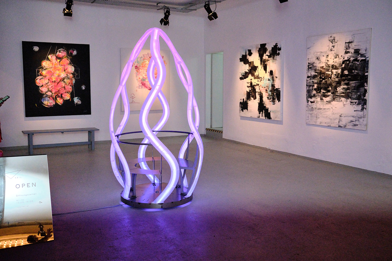 Eine zeitgenössische Kunstgalerie mit abstrakten Gemälden an weißen Wänden und einer zentralen Skulptur aus sich drehenden, leuchtenden violetten Lichtschläuchen. Eine Bank und ein Schild mit der Aufschrift "OPEN" sind ebenfalls zu sehen. Der Raum ist gut beleuchtet und modern.