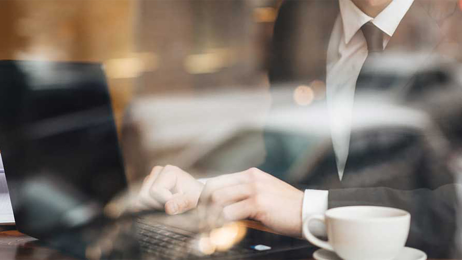Eine Person im Anzug tippt auf einem Laptop an einem Tisch mit einer Tasse Kaffee, gesehen durch ein Fenster mit Spiegelungen.
