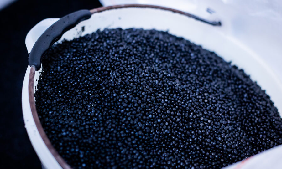 Ein weißer Behälter, der mit zahlreichen kleinen, runden, schwarzen Plastikkügelchen gefüllt ist, an deren Rand ein Stück schwarzes Material klebt.