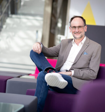 Ein Mann mit Brille, beigem Blazer, Jeans und weißen Turnschuhen sitzt entspannt auf einem modernen lila Sofa in einem hellen, modernen Innenraum und lächelt in die Kamera.