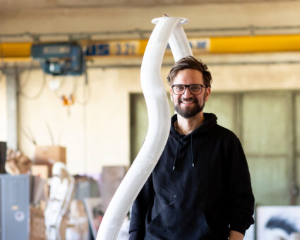Ein lächelnder Mann mit Brille und schwarzem Kapuzenpulli steht in einem Innenraum neben einer hohen, geschwungenen weißen Skulptur. Im Hintergrund sind Werkstattausrüstungen und -materialien bei weichem, natürlichem Licht zu sehen.