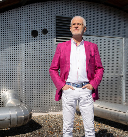 Ein älterer Mann mit weißem Bart steht selbstbewusst und mit den Händen in den Taschen in einem leuchtend rosa Blazer, einem weißen Hemd und einer weißen Hose vor einem modernen, metallischen Gebäude im Industriestil.