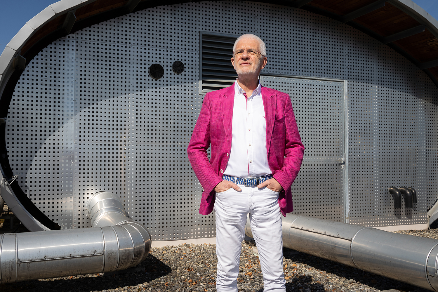 Ein älterer Mann mit weißem Bart steht selbstbewusst und mit den Händen in den Taschen in einem leuchtend rosa Blazer, einem weißen Hemd und einer weißen Hose vor einem modernen, metallischen Gebäude im Industriestil.