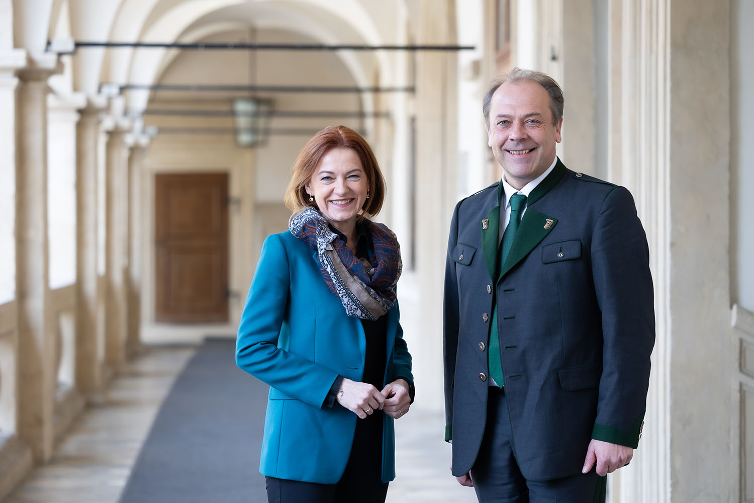 Zwei Personen stehen lächelnd in einem hellen, gewölbten Flur. Die Frau trägt eine blaugrüne Jacke und ein gemustertes Halstuch, der Mann einen traditionellen dunklen Anzug mit grünen Akzenten. Der Korridor ist mit Säulen und einer Laterne über dem Kopf versehen.