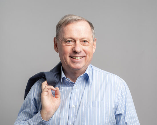 Ein älterer Mann mit kurzen grauen Haaren lächelt in die Kamera. Er trägt ein hellblau gestreiftes Hemd und hat eine dunkle Jacke über der Schulter.