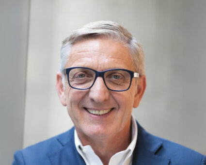 Ein lächelnder älterer Mann mit kurzen grauen Haaren, der eine dunkel umrandete Brille, ein weißes Hemd und eine blaue Anzugsjacke trägt. Der Hintergrund ist unscharf und neutral.