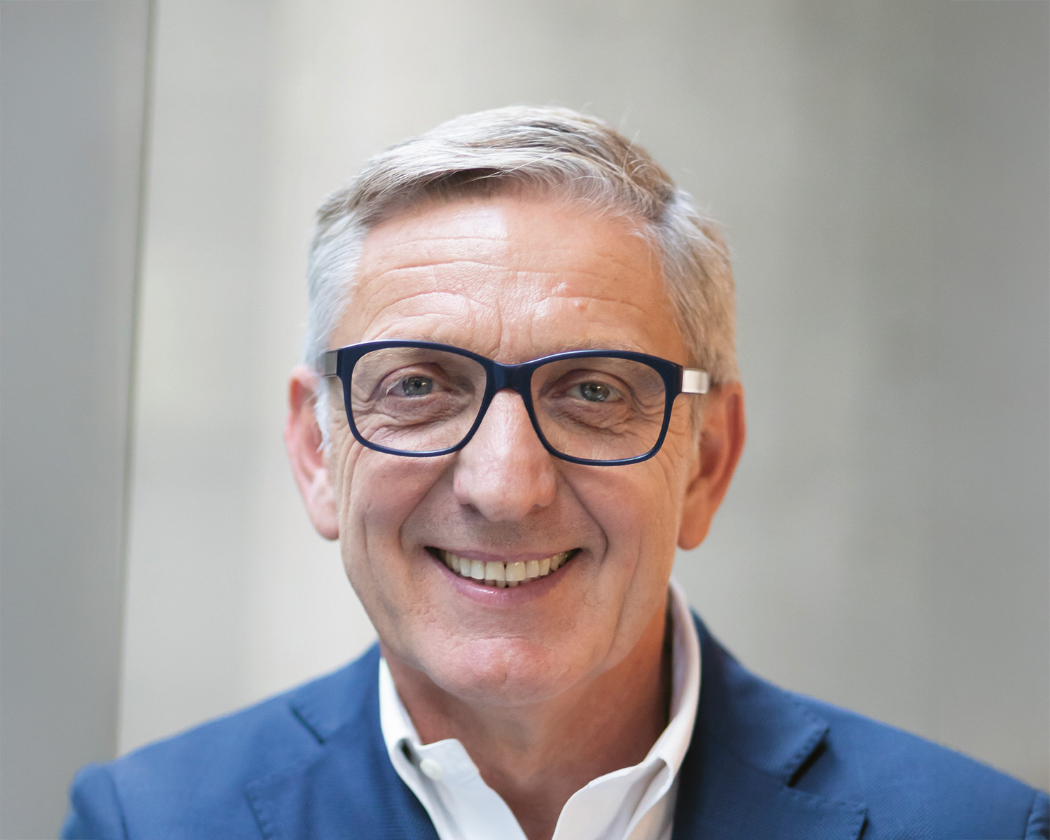 Ein lächelnder älterer Mann mit kurzen grauen Haaren, der eine dunkel umrandete Brille, ein weißes Hemd und eine blaue Anzugsjacke trägt. Der Hintergrund ist unscharf und neutral.